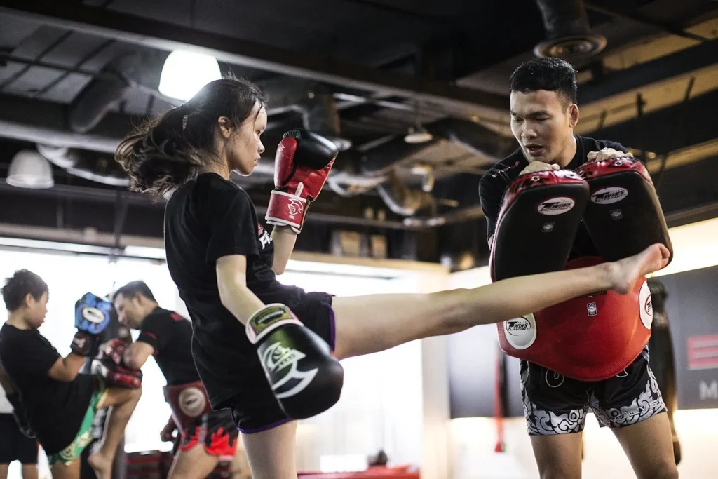 Instructor de Muay Thai sosteniendo protectores mientras una estudiante practica la técnica de patadas altas en un gimnasio